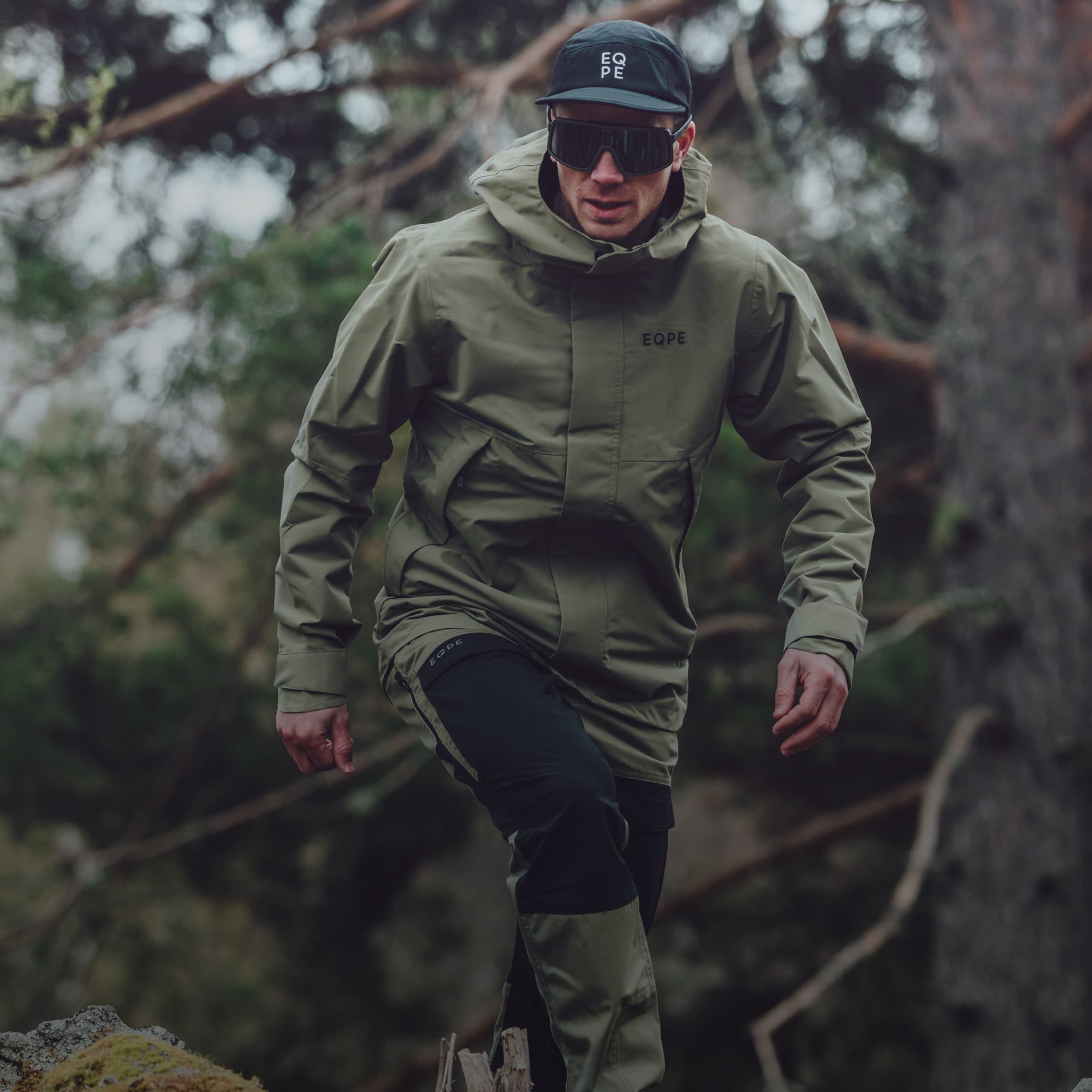 Person in green outdoor gear and sunglasses, walking in a forest, wearing a cap with "EQPE" logo.