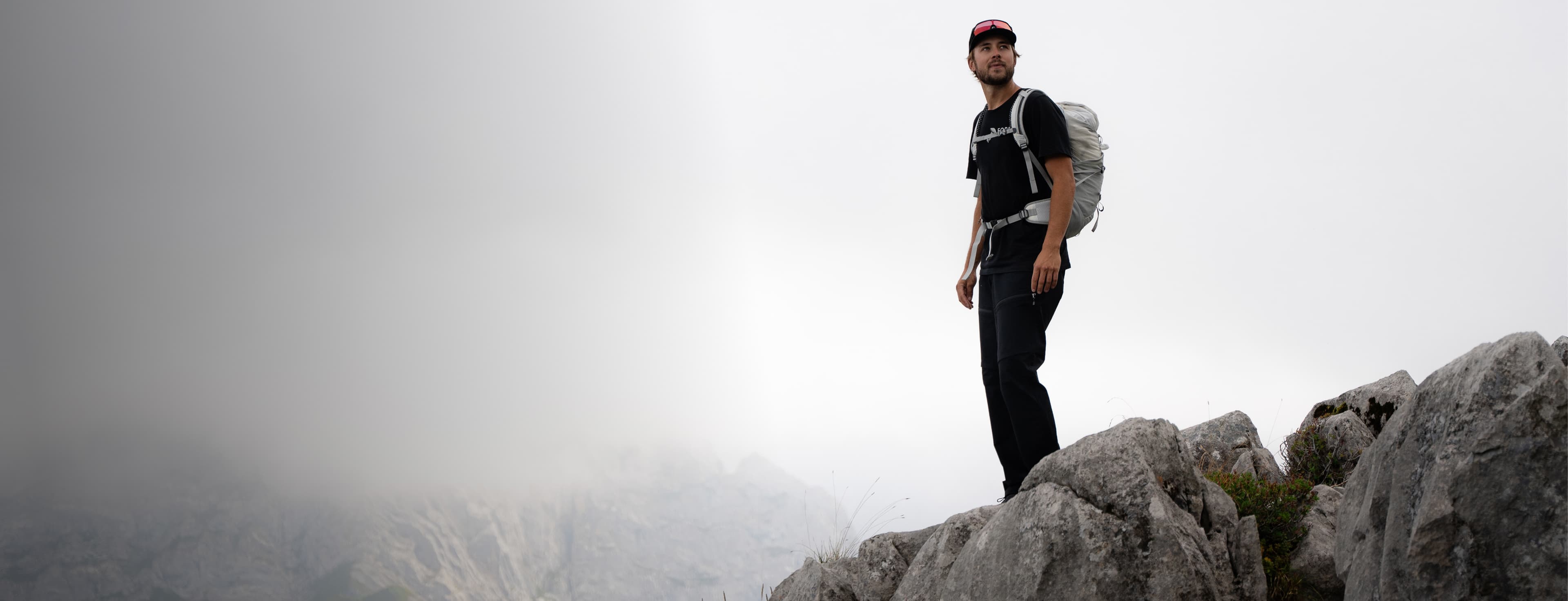 A person stands on a rocky mountain peak with a backpack, looking into the distance. The background is misty and overcast.