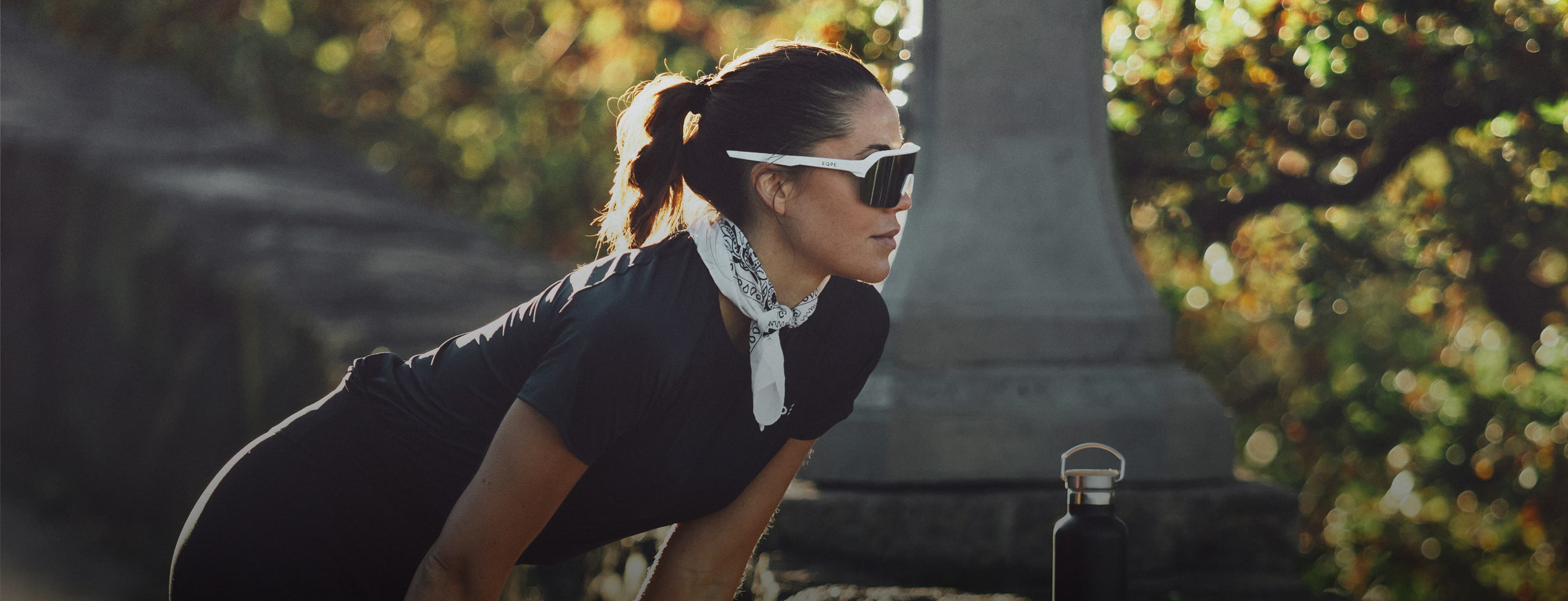 Woman in athletic wear and sunglasses leans forward outdoors, with trees and sunlight in the background, and a water bottle nearby.