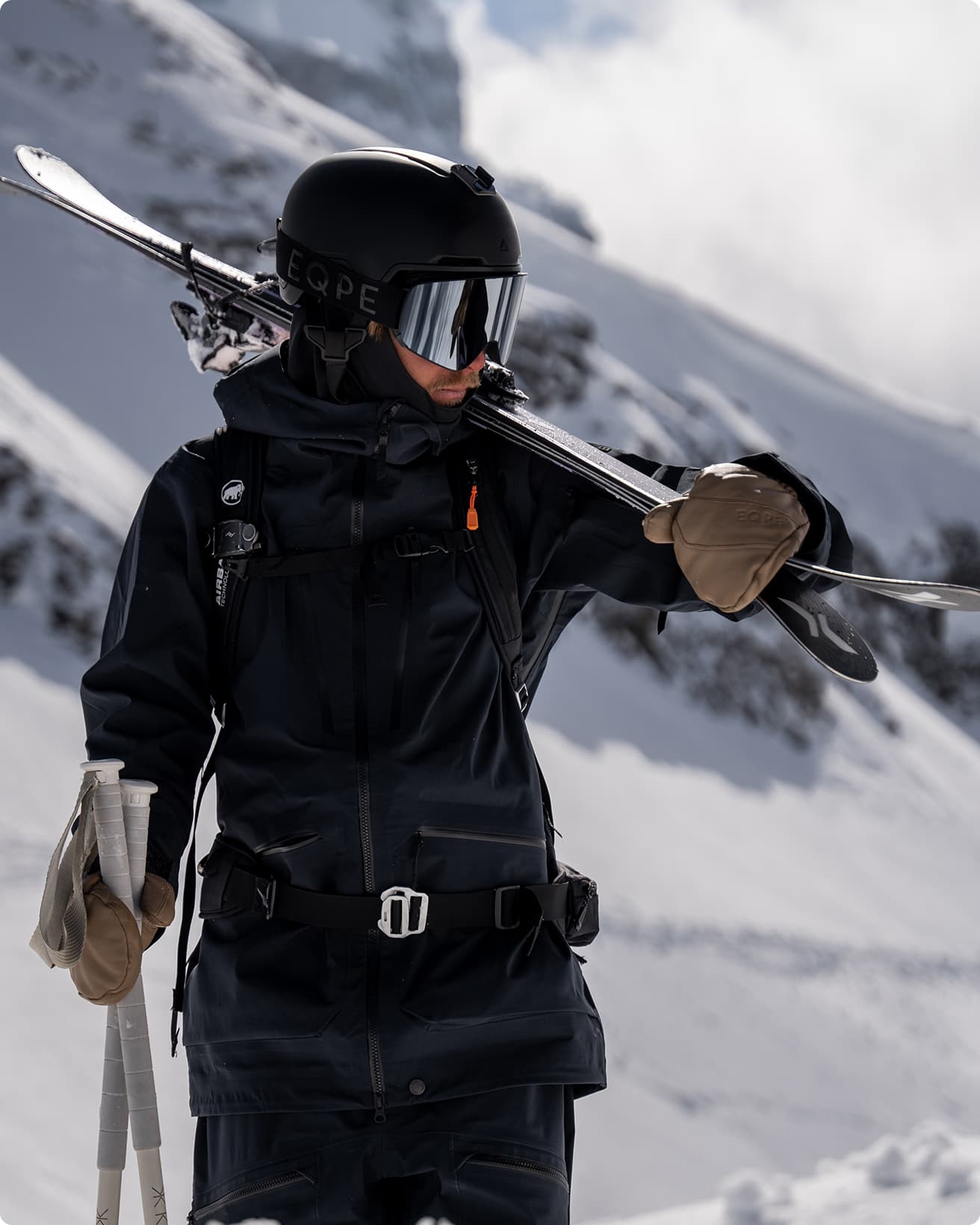 Person in dark ski gear holding skis over shoulder, standing on snowy mountain with white skies in the background.