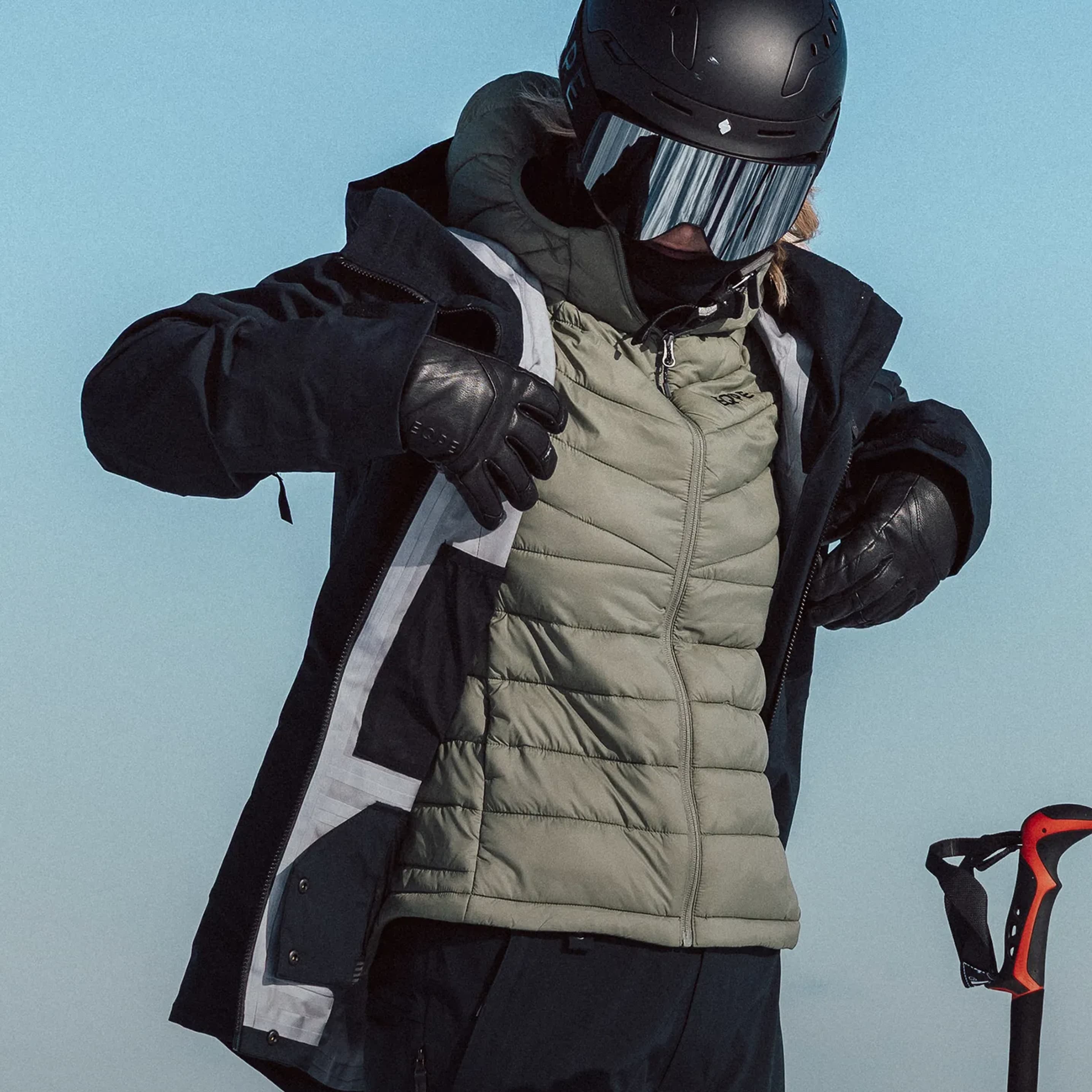 A person wearing winter gear, including a helmet, goggles, and gloves, adjusts their jacket, standing next to a ski pole against a clear sky.
