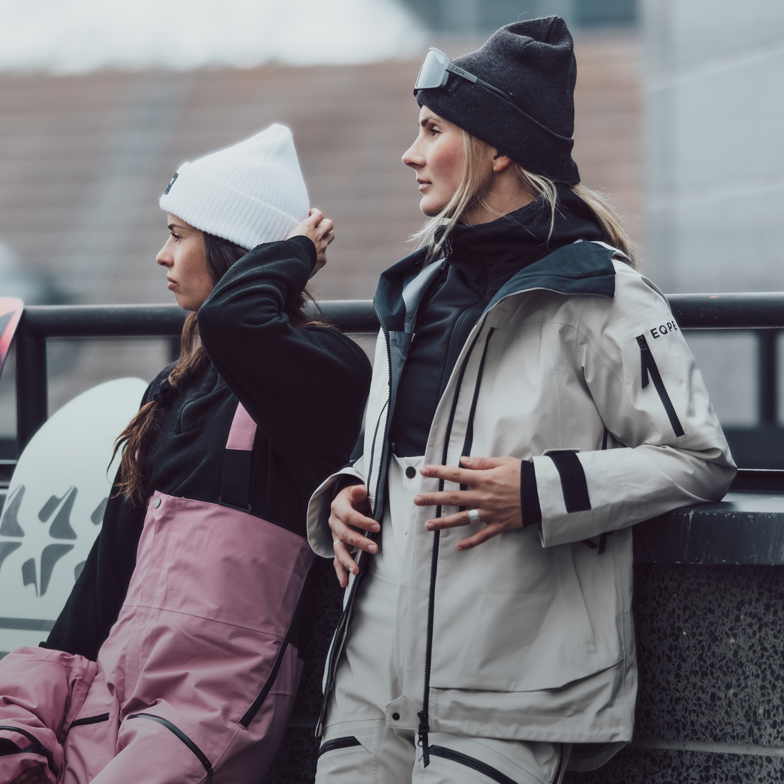 Two women in ski gear stand outdoors. Snowboards in the background.