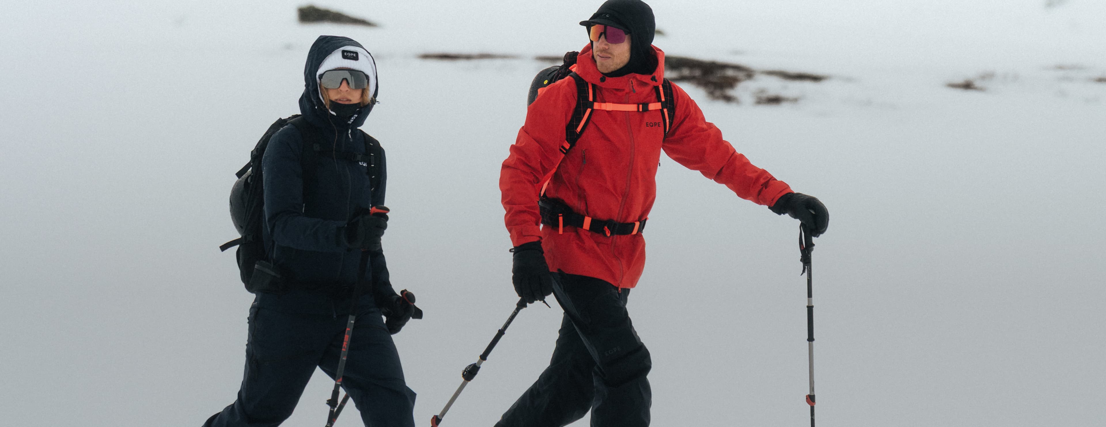 Two people in winter gear—one wearing a red jacket—trekking with poles across a snowy landscape.
