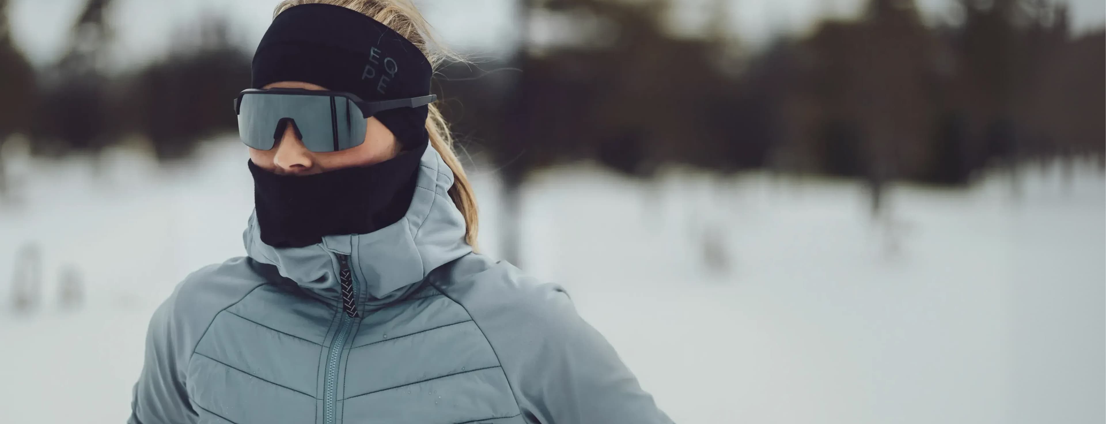 Person dressed in winter gear, including sunglasses and a headband, stands in a snowy landscape with blurred trees in the background.