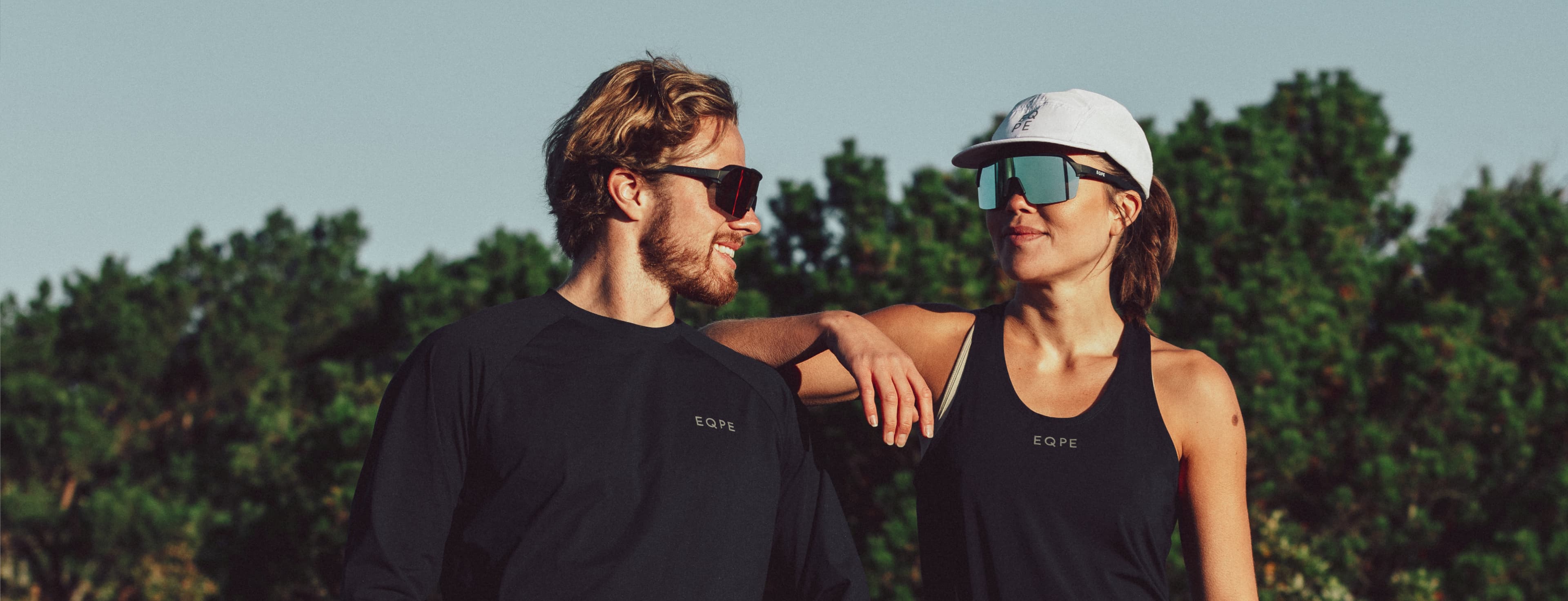 A man and woman in sports attire and sunglasses stand outdoors, smiling at each other. Trees are visible in the background.